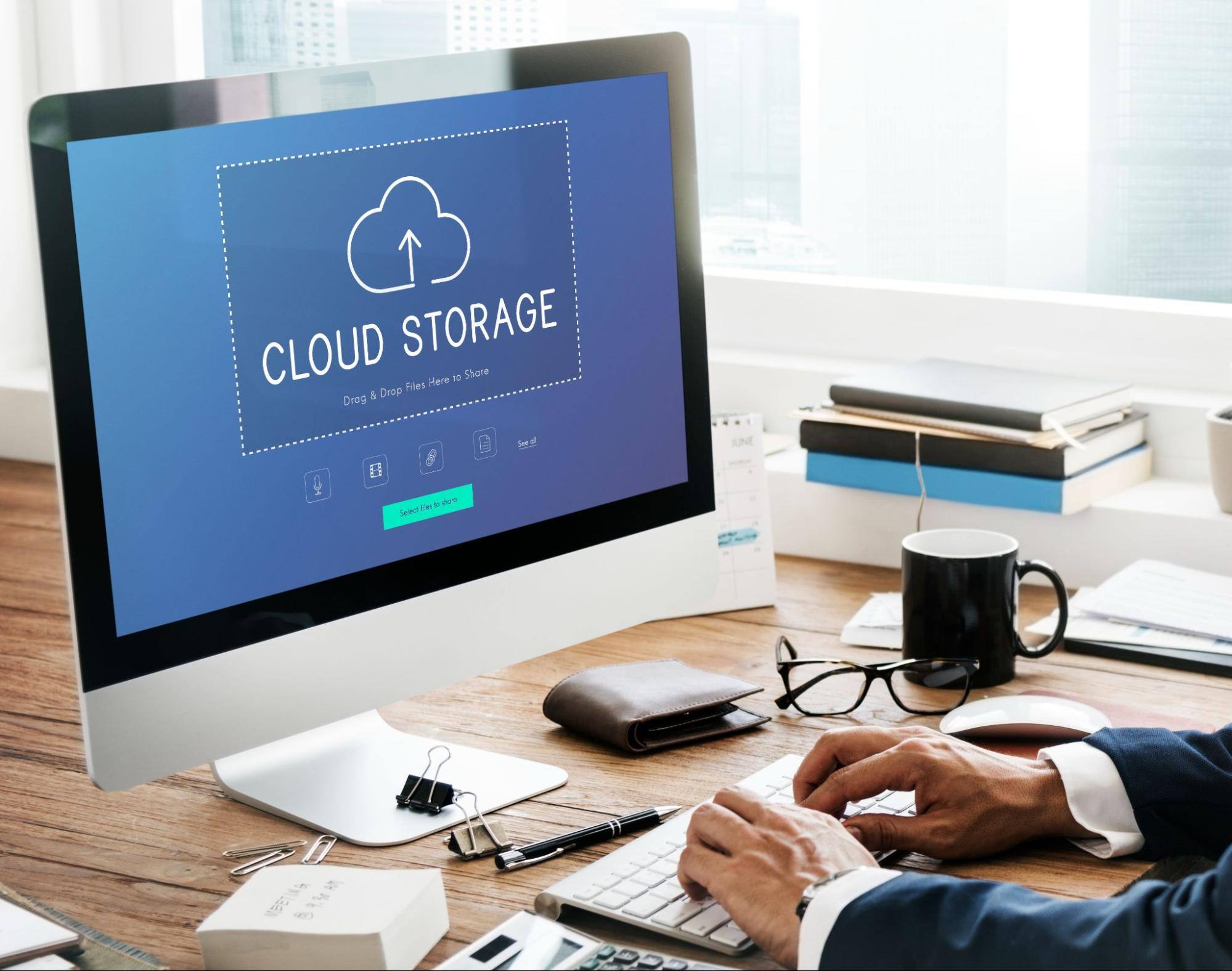 An unrecognized businessman works at a busy desk. His computer screen shows 'Cloud Storage' and an icon for data upload, representing cloud-based data management.